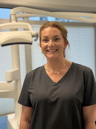 Kvinne i svart legefrakk smiler, står foran dentalutstyr i lyst rom.