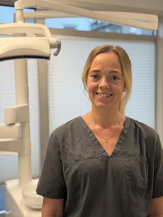 Kvinne med smil iført grå arbeidsklær, stående foran dentalutstyr i lyst rom.