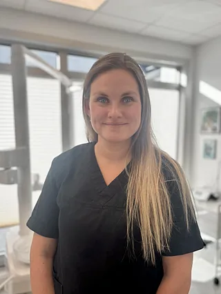 En kvinne med langt hår i en svart uniform står smilende i et lyst rom.